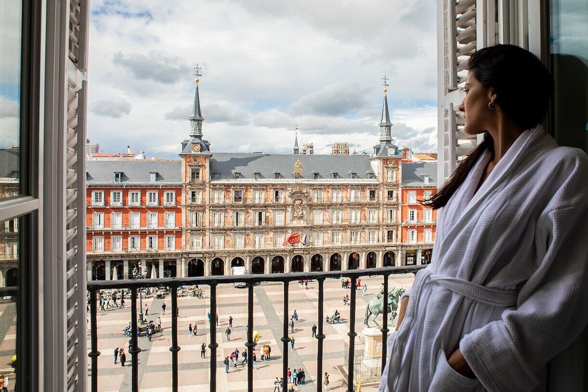 Une personne vêtue d’une robe blanche se tient près d’une fenêtre donnant sur une place historique, où se trouve un bâtiment remarquable, doté de deux tours et d’une statue.