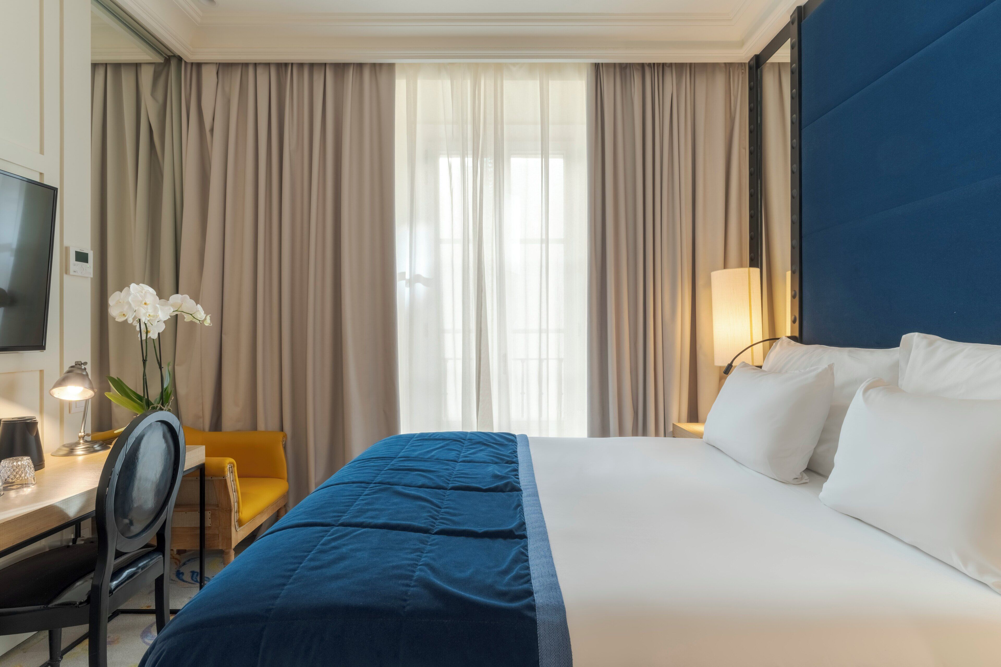 A hotel room with a bed, a desk, a chair, a TV, and a window with curtains.