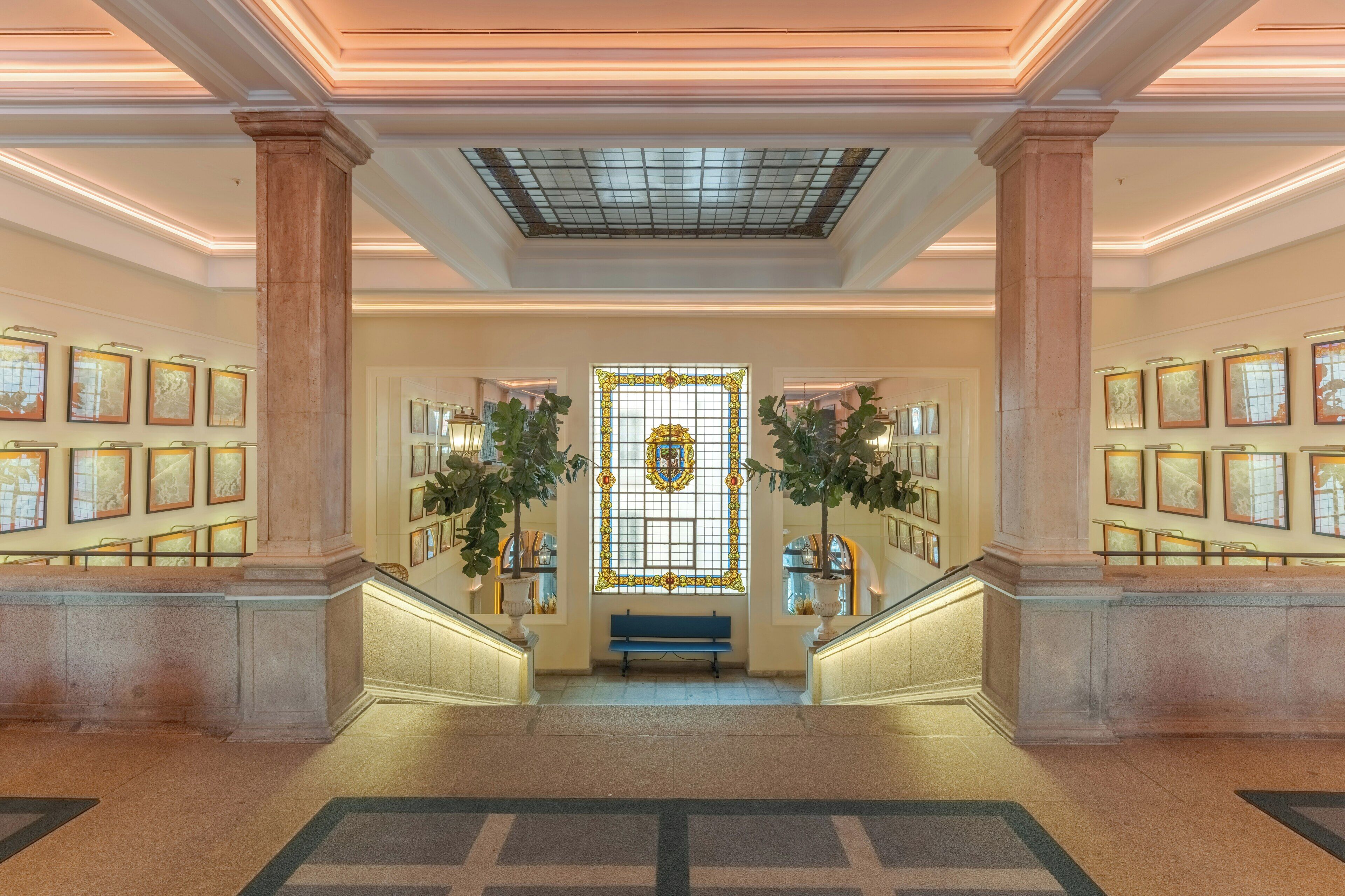 A grand lobby with a central staircase, framed artwork on the walls, and a decorative glass door.