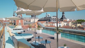 Area piscina sul tetto con poltrona a sdraio e ombrelloni, con vista su un paesaggio urbano ricco di edifici storici.