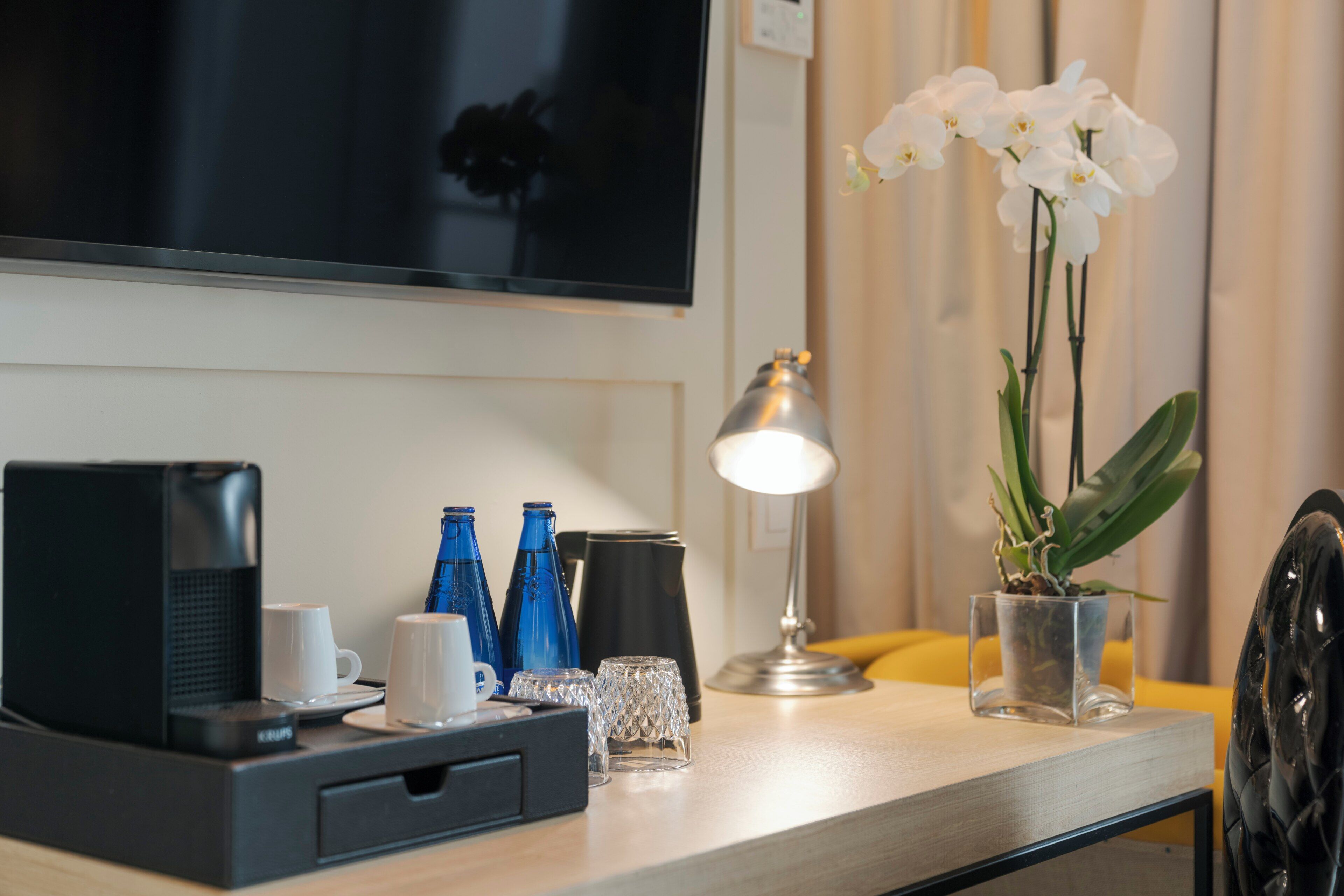 A hotel room with a flat-screen TV mounted on the wall, a desk with a coffee maker, cups, and a lamp. There are potted orchids and a small plant on the desk.