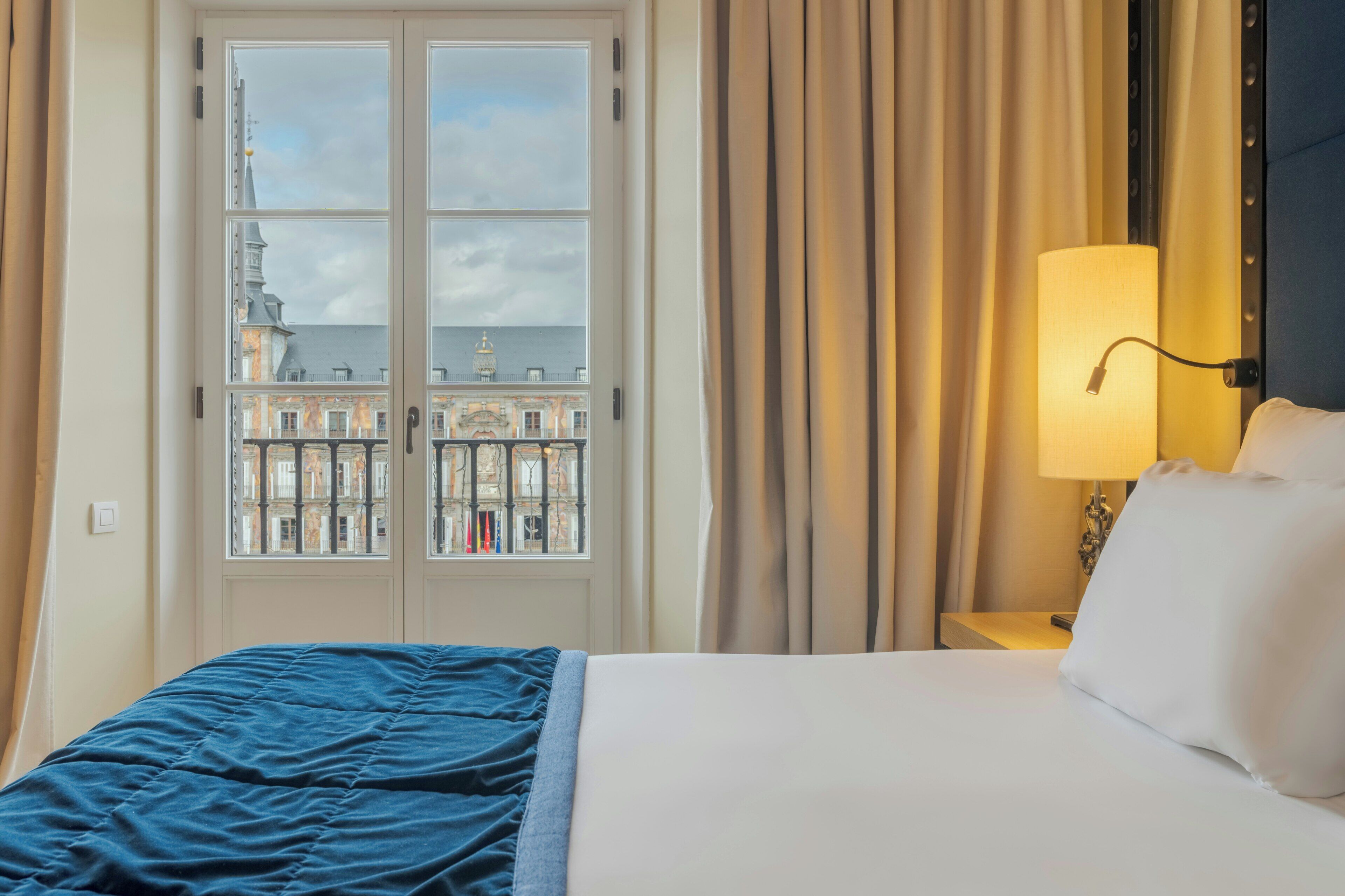 A hotel room with a bed, a nightstand, a lamp, and a view of buildings through a window.