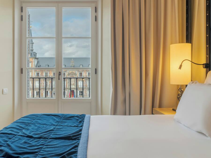 Habitación de hotel con cama, mesita de noche, lámpara y vistas a edificios a través de una ventana.