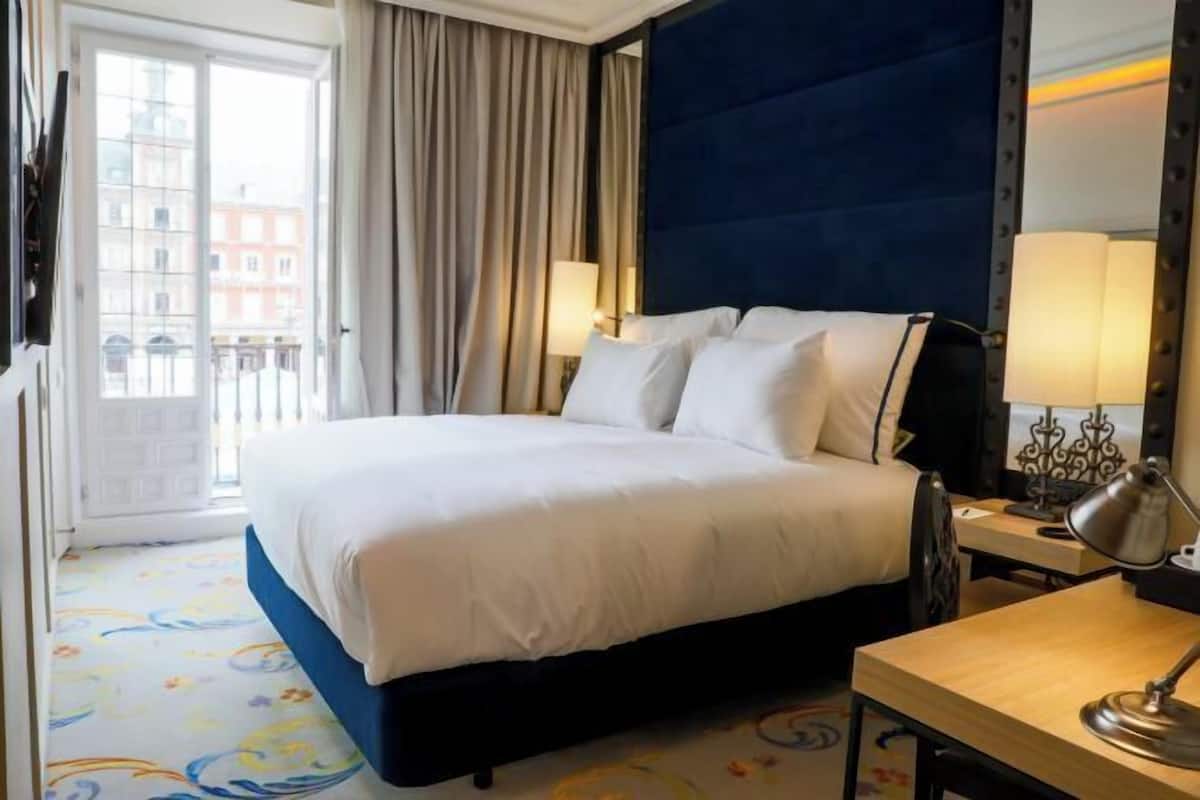 Une chambre d’hôtel avec un lit, un bureau, une lampe, une fenêtre avec des rideaux et une vue sur les bâtiments environnants.