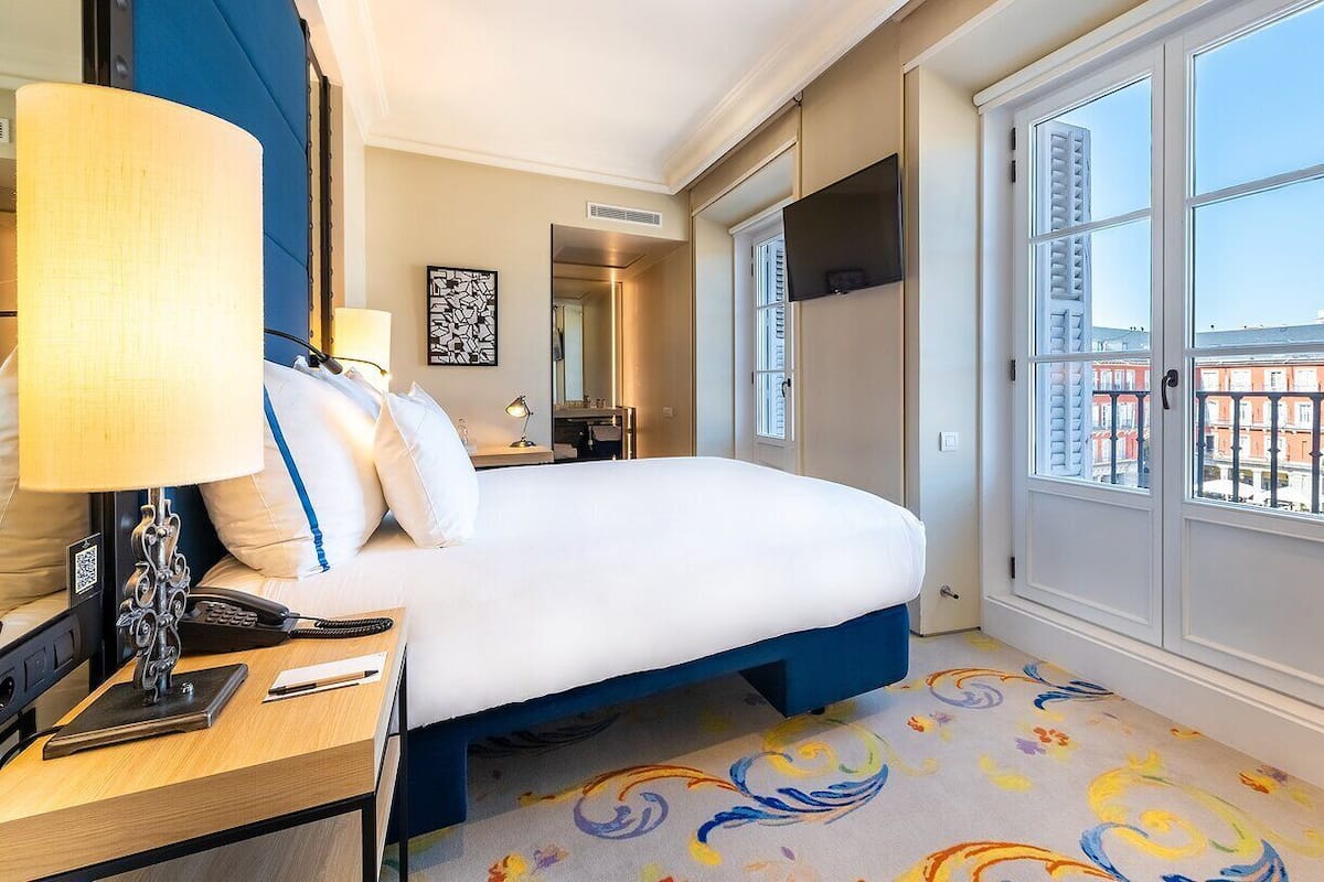 Une chambre d’hôtel avec un lit, une table de chevet avec une lampe, un téléphone et une fenêtre donnant sur des immeubles.