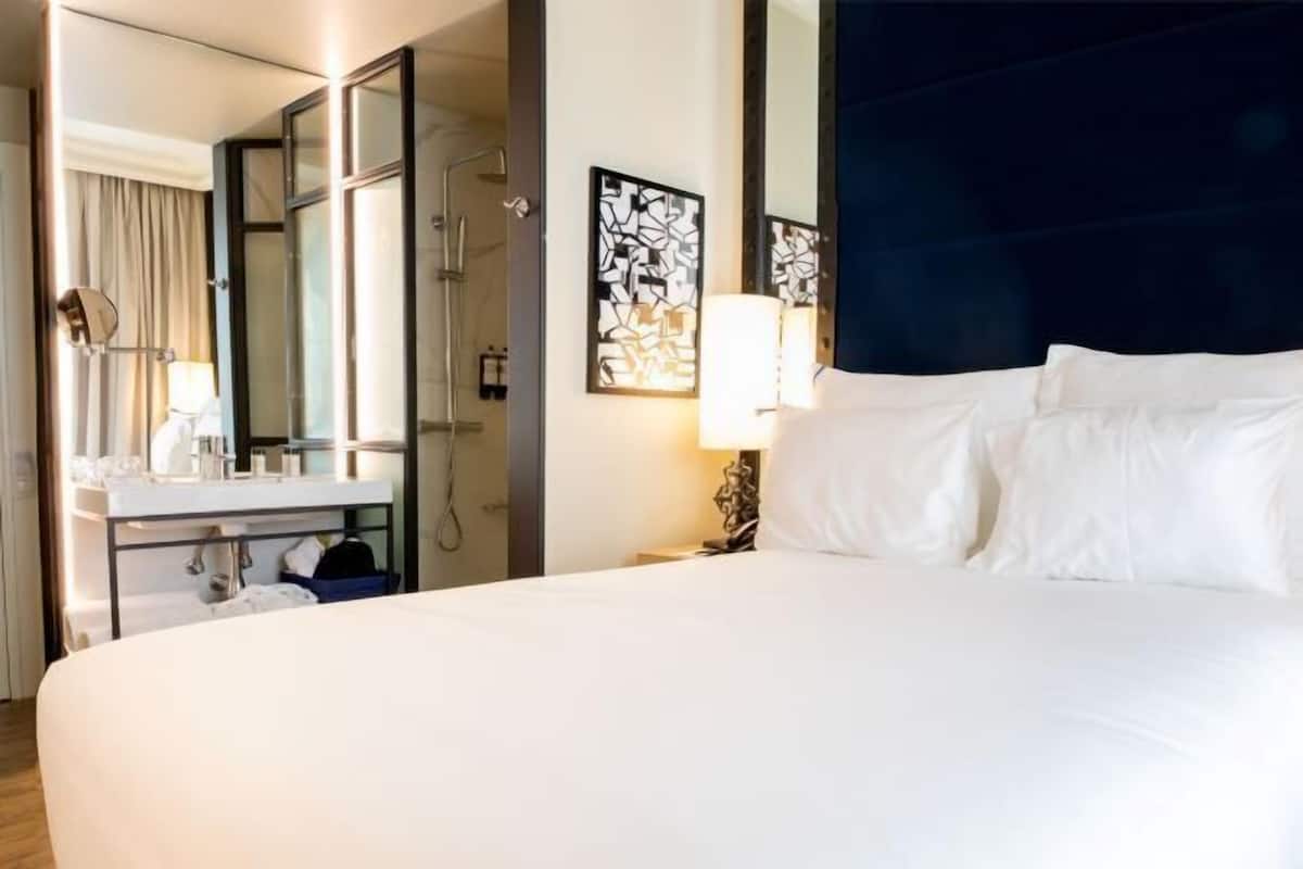Une chambre d’hôtel avec un grand lit, une salle de bain avec un miroir et un lavabo, et un luminaire fixé au mur.