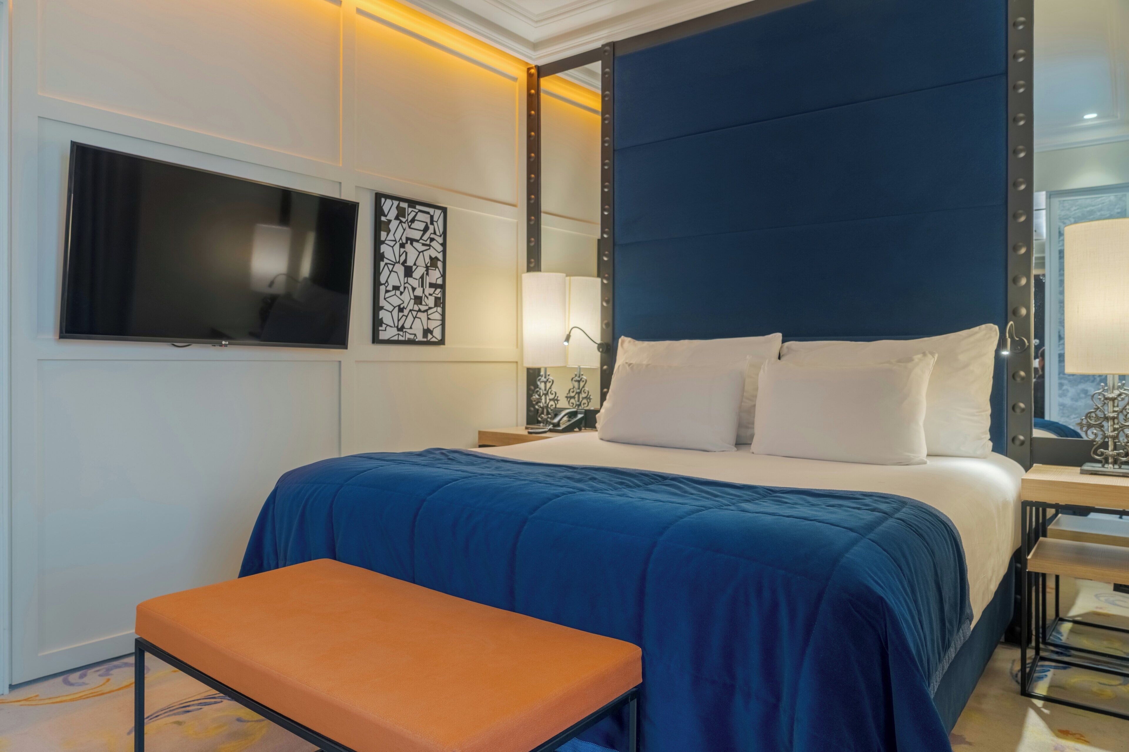 Habitación de hotel con una cama grande, un televisor de pantalla plana montado en la pared, una mesita de noche con lámpara y un banco.