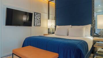 Habitación de hotel con una cama grande, un televisor de pantalla plana montado en la pared, una mesita de noche con lámpara y un banco.