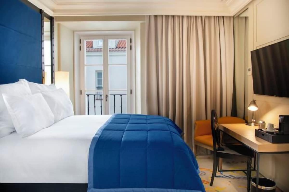 A hotel room with a bed, a desk, a chair, a lamp, and a view of the outside.