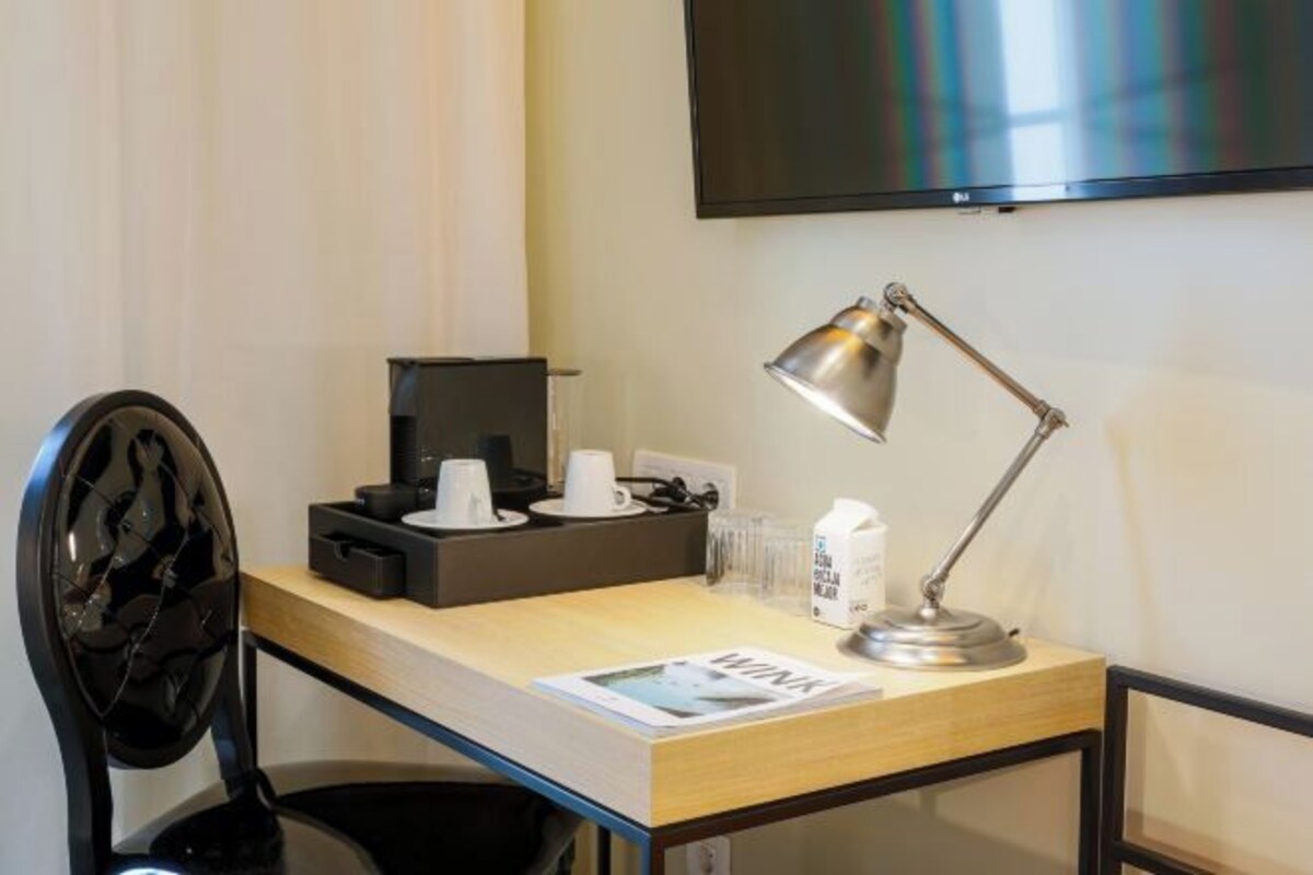 A hotel room with a desk, a chair, a TV mounted on the wall, a coffee maker, and a lamp.