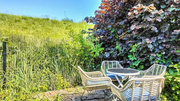 Une terrasse de jardin aménagée avec des meubles en osier et une table, entourée d’une végétation luxuriante et d’une clôture en métal.
