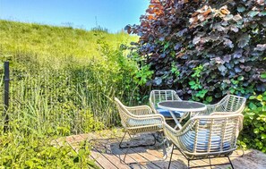 Une terrasse de jardin aménagée avec des meubles en osier et une table, entourée d’une végétation luxuriante et d’une clôture en métal.