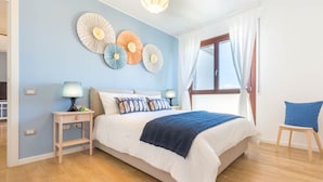 A bedroom with a bed, bedside tables, a lamp, decorative wall art, a window with curtains, and a chair. - Appartamento 5th Floor (Cagliari)
