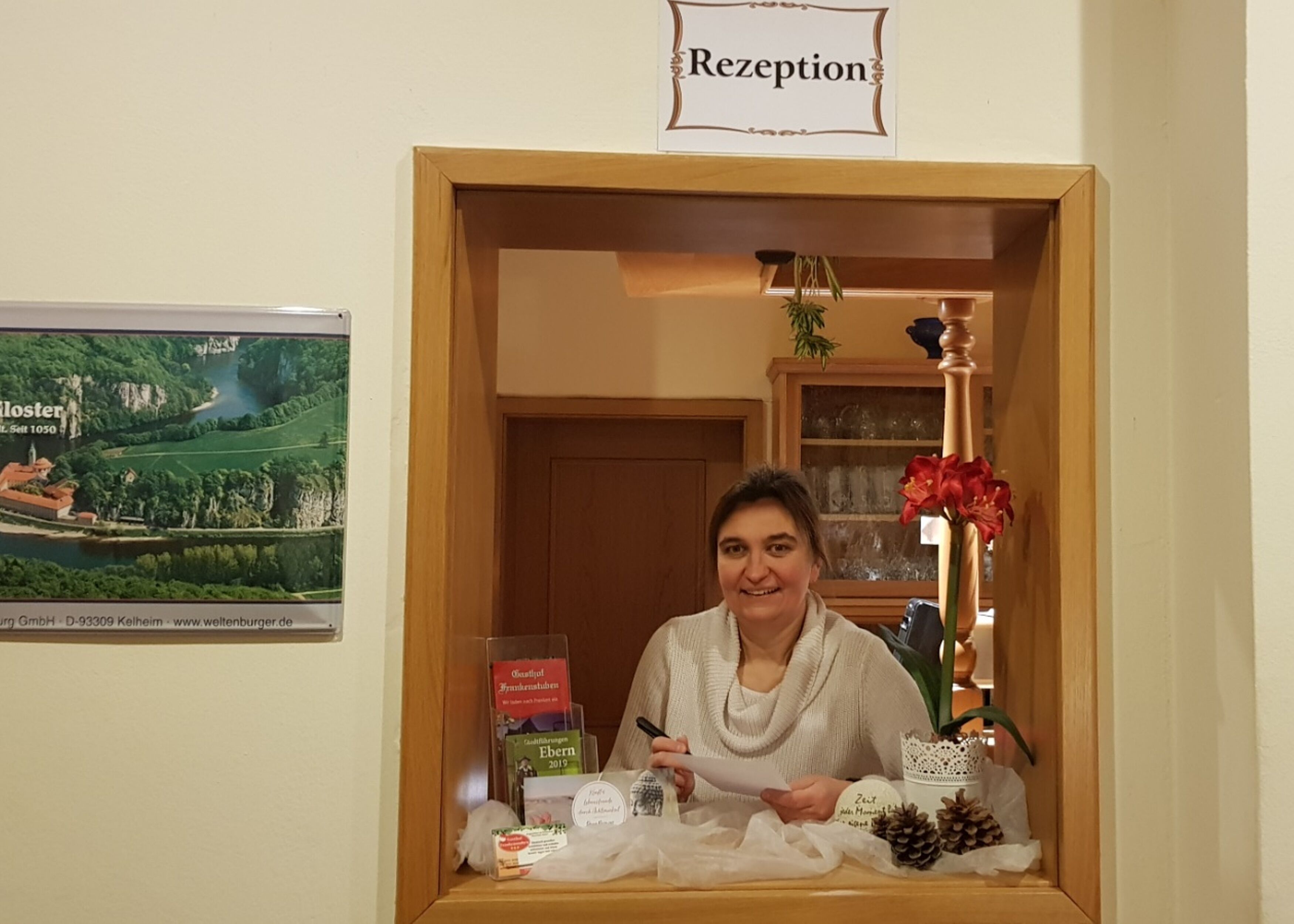 Un mostrador de recepción con un cartel encima, un cuadro en la pared y una pequeña mesa con una planta y un libro.
