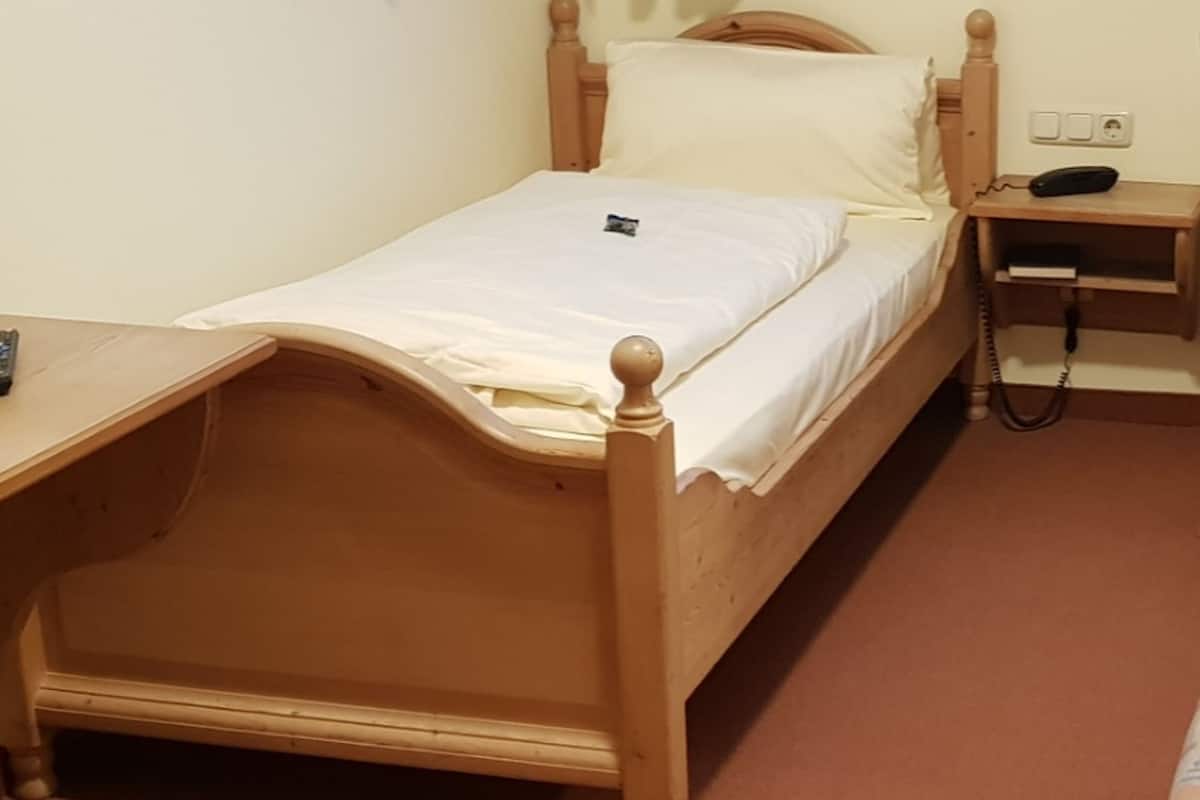 A single bed with a wooden frame, a bedside table with a phone, and a power outlet on the wall.