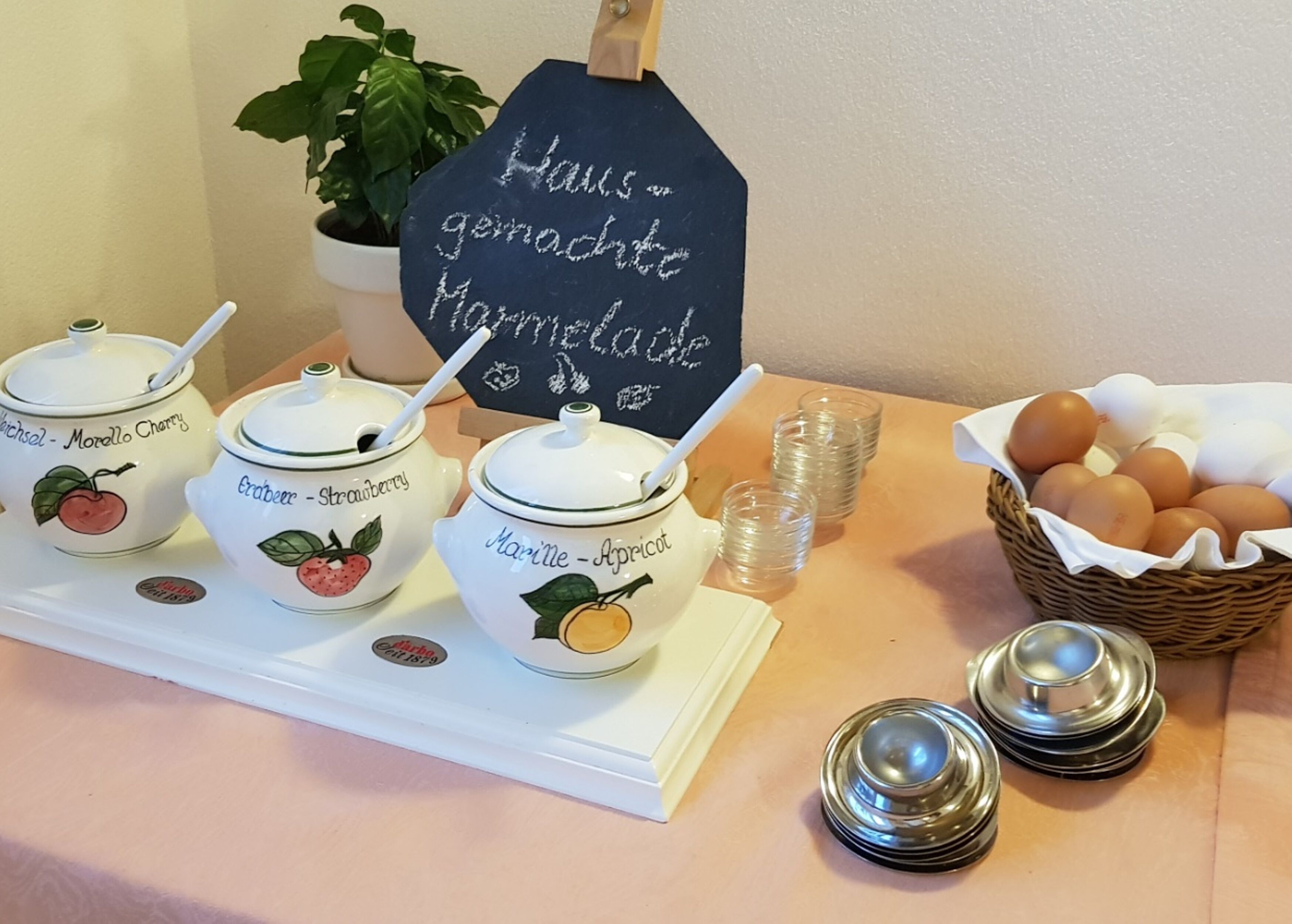 Una presentación para el desayuno con frascos etiquetados con nombres de frutas, un letrero de pizarra, una cesta de huevos y una planta en maceta.