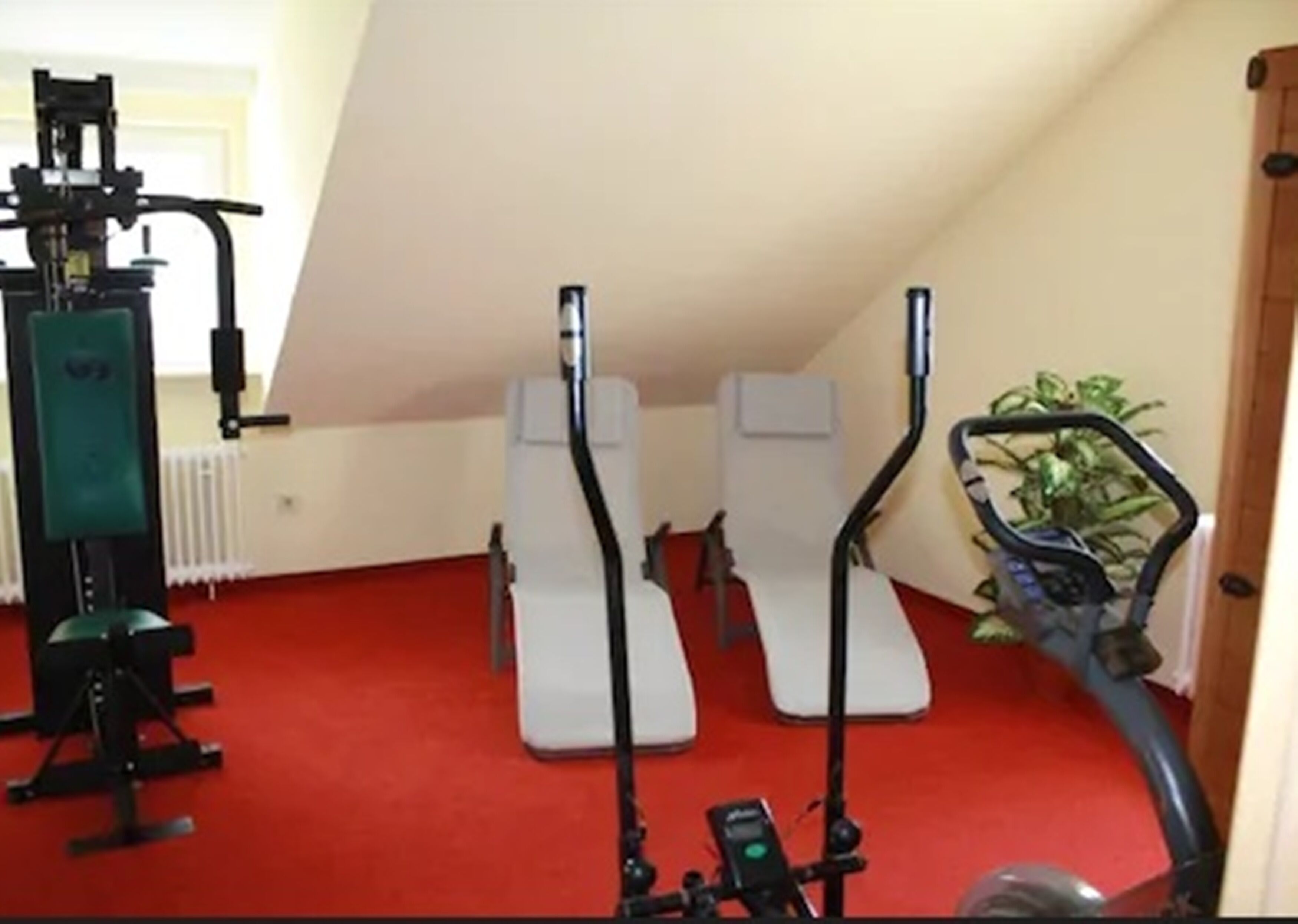 A room with a red carpet, two white exercise chairs, a green exercise machine, and a blue vacuum cleaner.