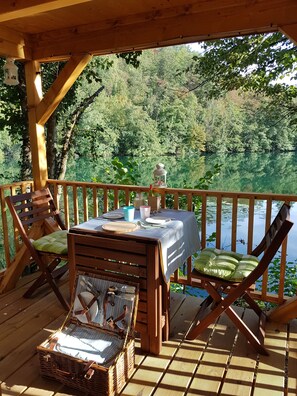 Una terraza de madera con una mesa dispuesta para dos, con vista a un lago rodeado de árboles.