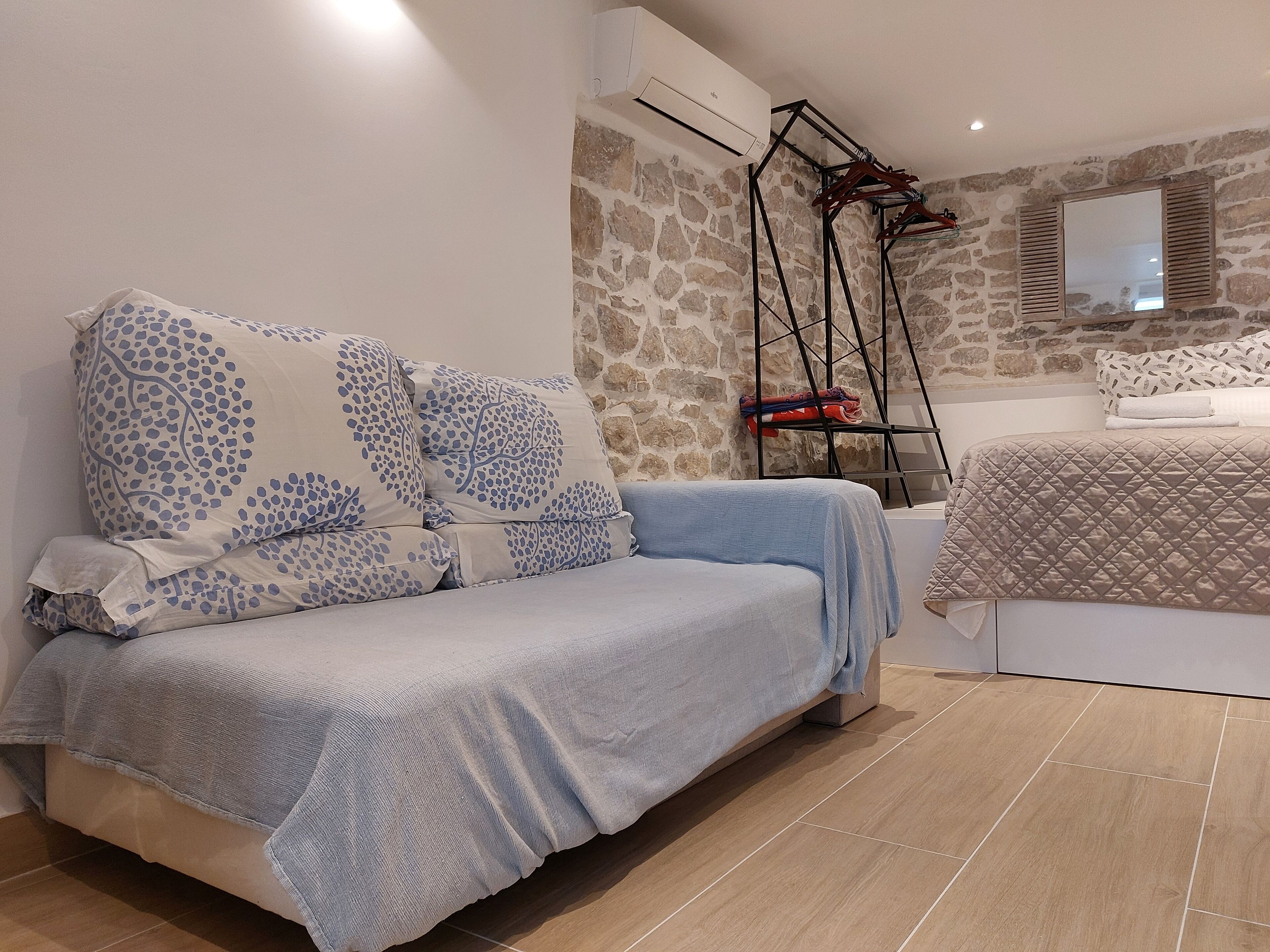 A bedroom with a stone wall, a bed with a quilted cover, a sofa with patterned cushions, and a hanging clothes rack.