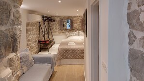 Ein schmaler Flur mit einer Steinsäule an einer Seite und einem Bett an der anderen. Eine Holzleiter lehnt an der Wand, und ein Fenster mit Fensterläden ist zu sehen.