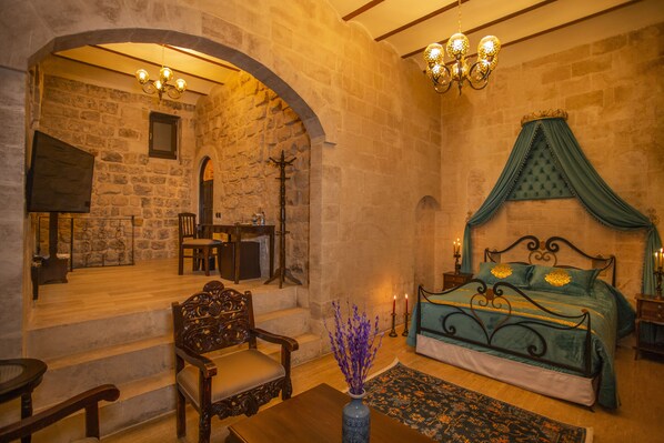 Quarto com parede de pedra, cama com dossel verde, cadeira de madeira, mesa e vaso com flores.