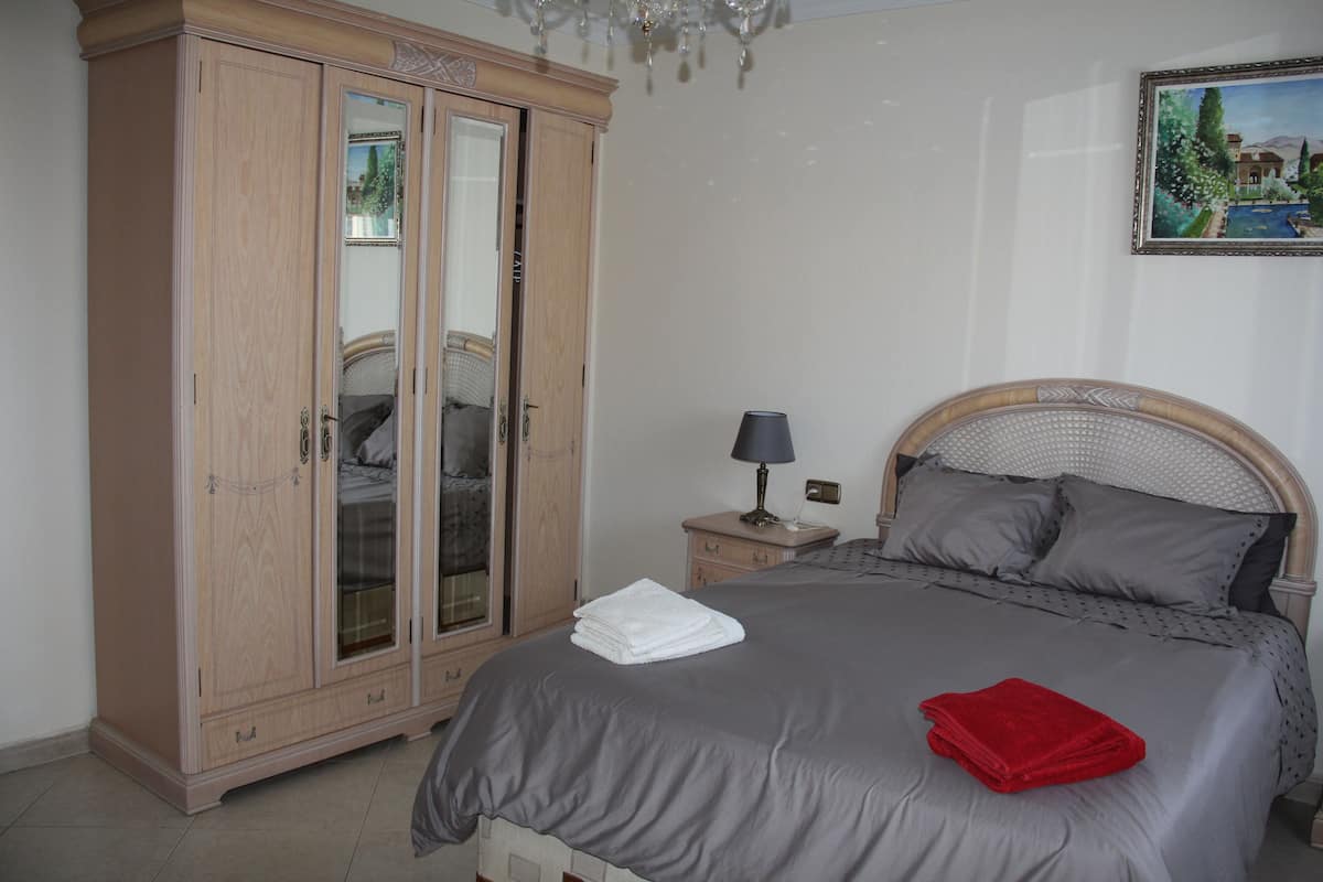 A bedroom with a large wardrobe, a bed with grey bedding, a nightstand with a lamp, and a painting on the wall.