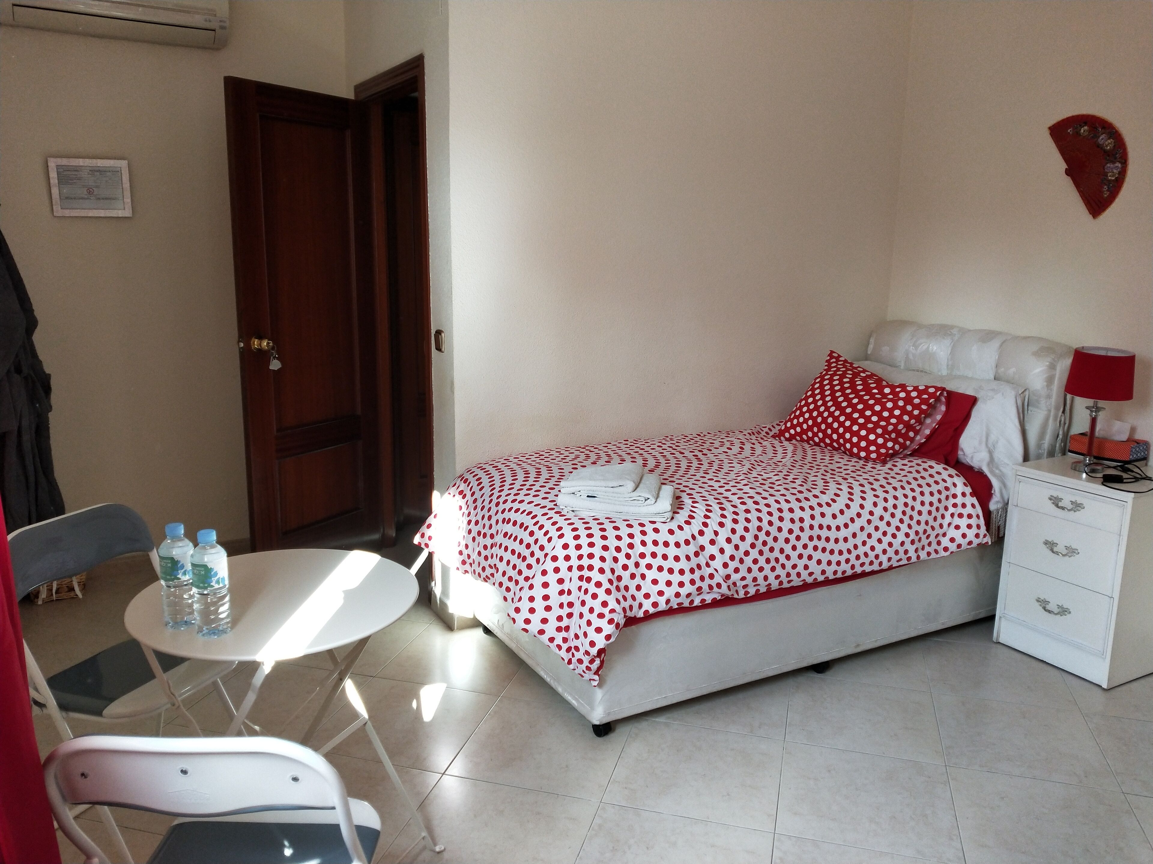 Une chambre bien rangée, avec un lit, une table de chevet et une petite table sur laquelle sont posées des bouteilles d’eau.