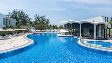 A large, clear blue pool with lounge chairs and umbrellas, surrounded by palm trees and modern buildings.