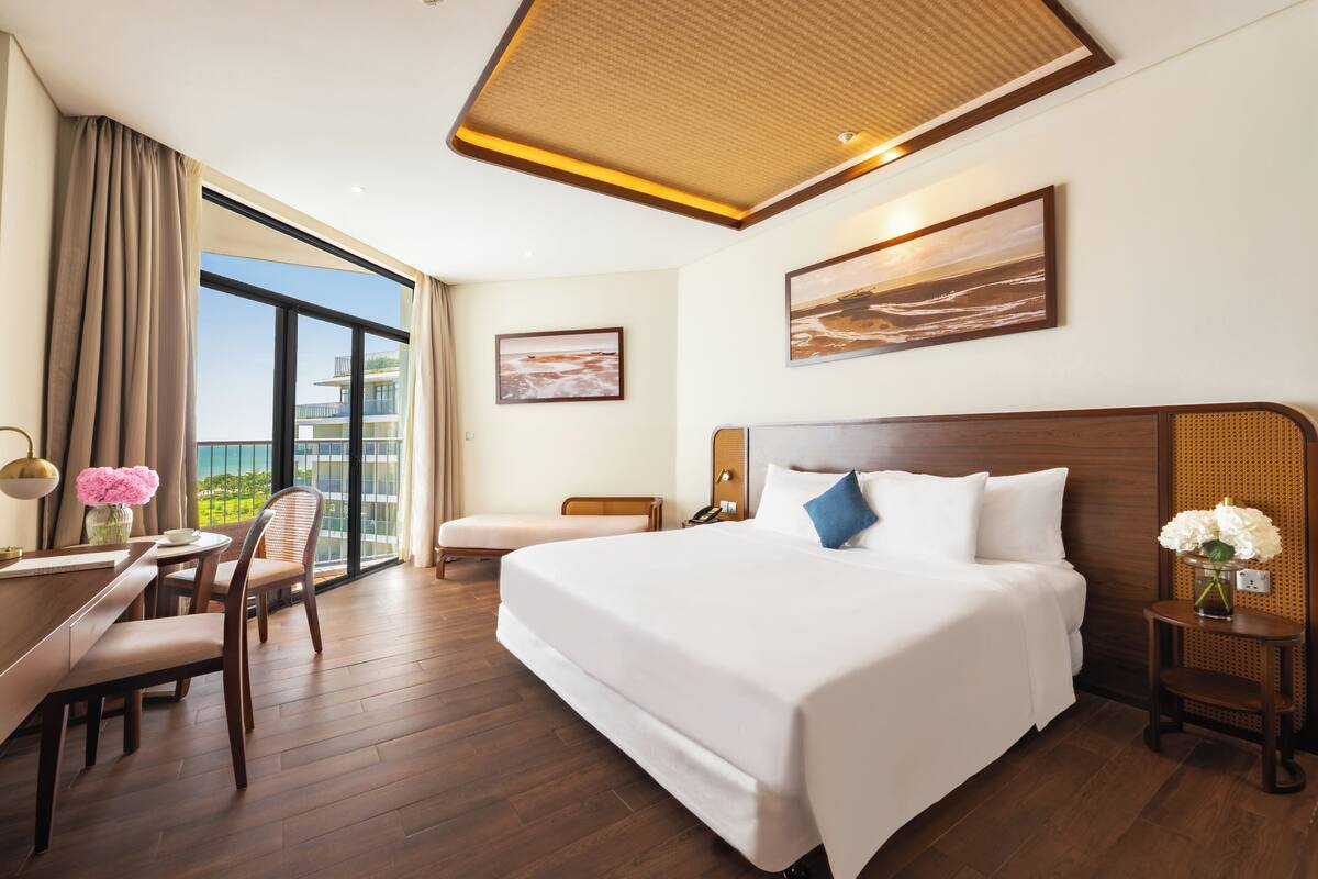 Ein Hotelzimmer mit einem großen Bett, einem Schreibtisch mit Stuhl, einem kleinen Tisch mit Blumenvase und Blick ins Freie.