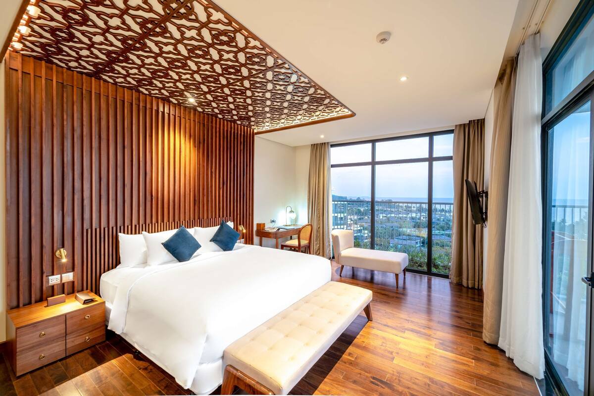Ein modernes Hotelzimmer mit einem großen Bett, einem Holz-Kopfteil, einer Bank, einem Schreibtisch und Blick auf die Stadt durch große Fenster.
