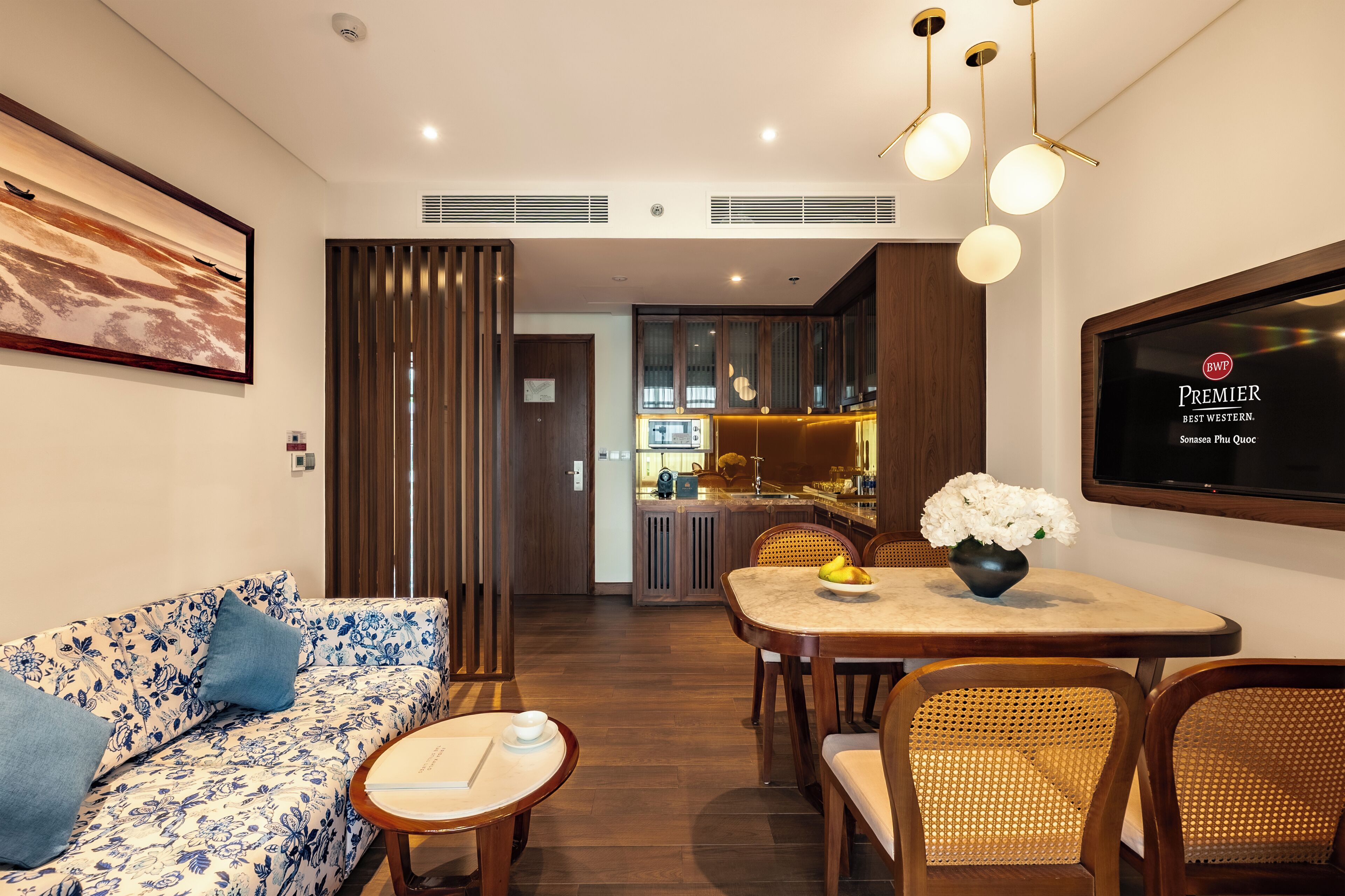 Una sala moderna con sofá, mesa de comedor y sillas, un televisor montado en la pared y una cocina al fondo.