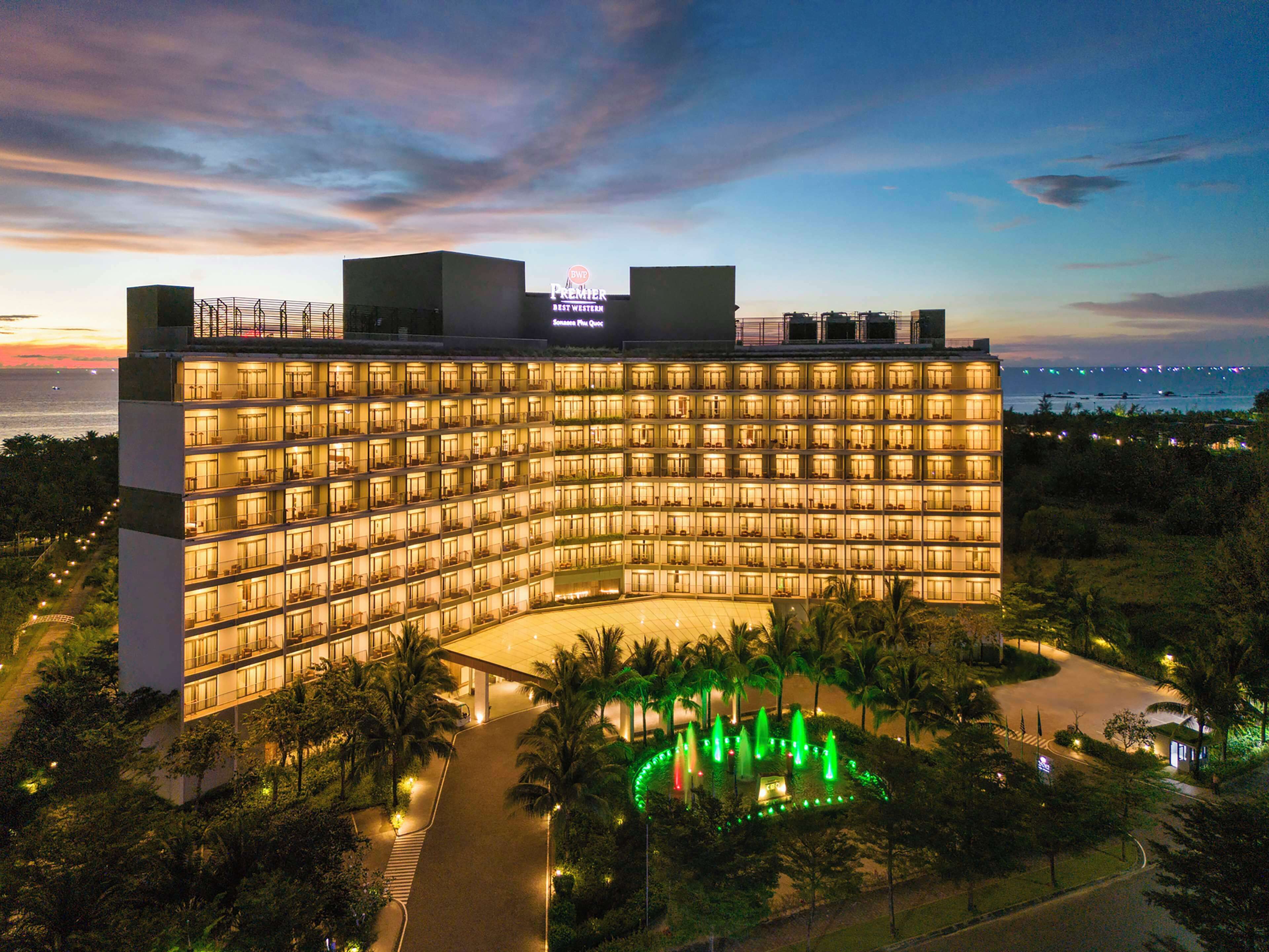 A large, multi-story hotel with a curved facade, illuminated windows, and a landscaped garden with fountains.