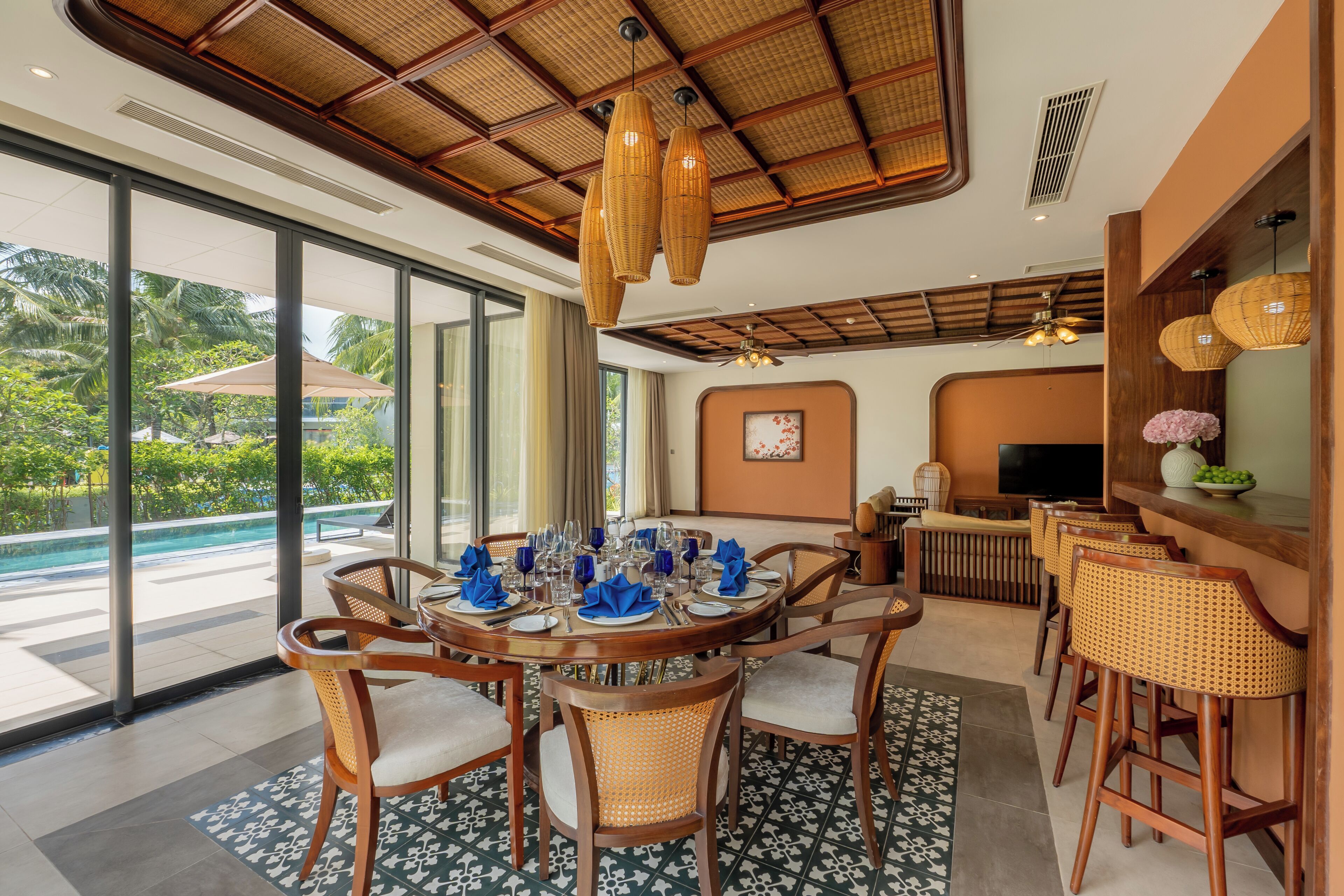 A dining area with a round table set for six, surrounded by wicker chairs, a pool view, and a bar area with wicker chairs and a TV.