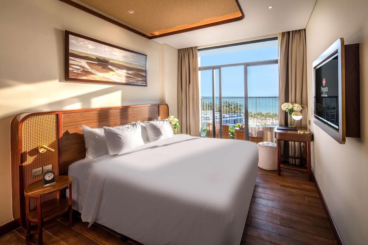 Ein Hotelzimmer mit einem großen Bett, einem Fernseher, einem Fenster mit Meerblick und einem kleinen Tisch mit Blumen.