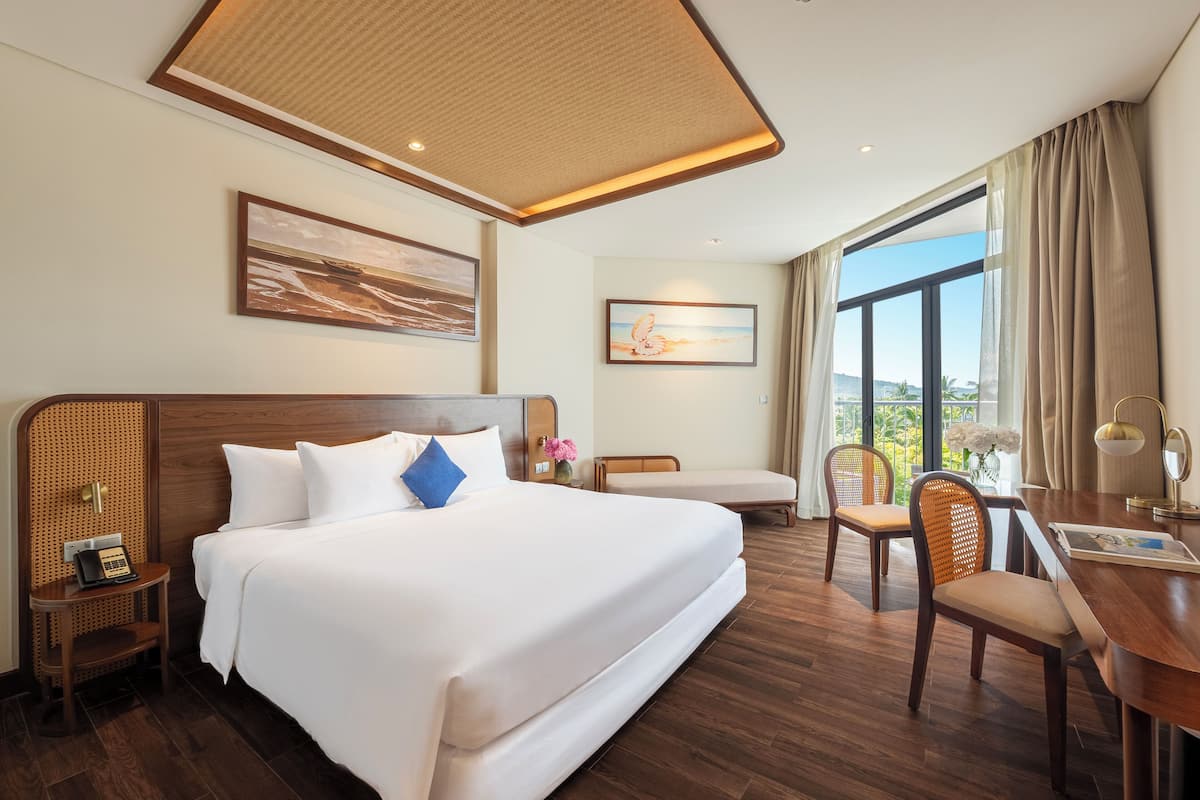 Ein Hotelzimmer mit einem großen Bett, einem Schreibtisch und einem Stuhl. An der Wand hängen zwei Bilder, ein Fenster mit Vorhängen und eine Deckenleuchte.