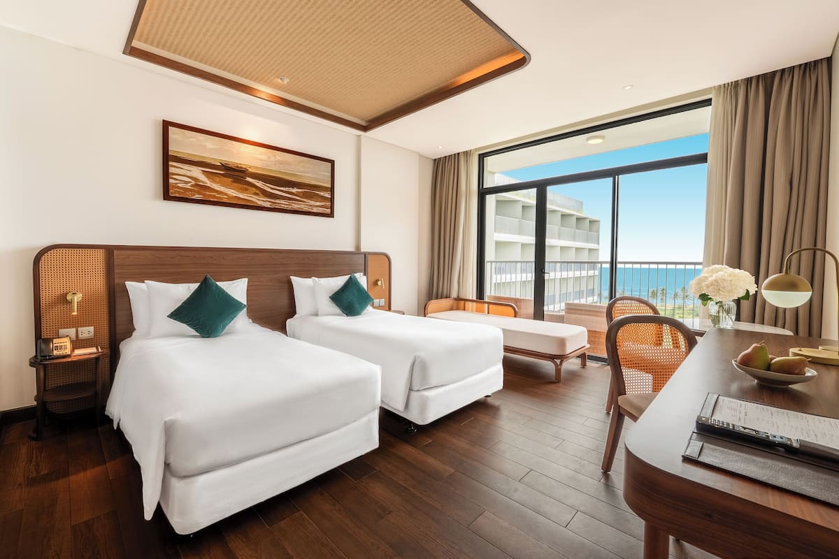 Ein Hotelzimmer mit zwei Betten, einem Esstisch, einem Stuhl, einem kleinen Tisch mit einer Lampe und einem großen Fenster mit Blick auf das Meer und Gebäude.