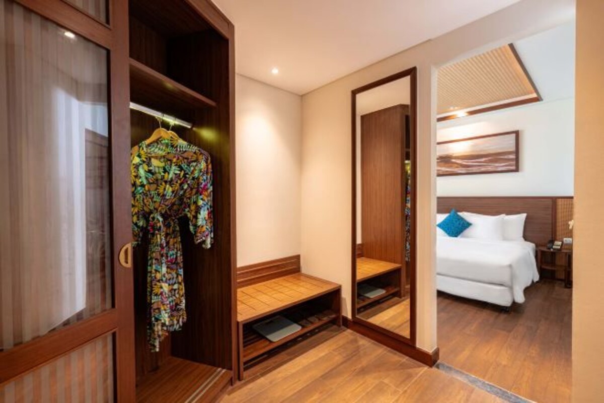 Une chambre d’hôtel avec une armoire en bois, un lit avec une literie blanche et un oreiller bleu, et un miroir qui reflète la pièce.