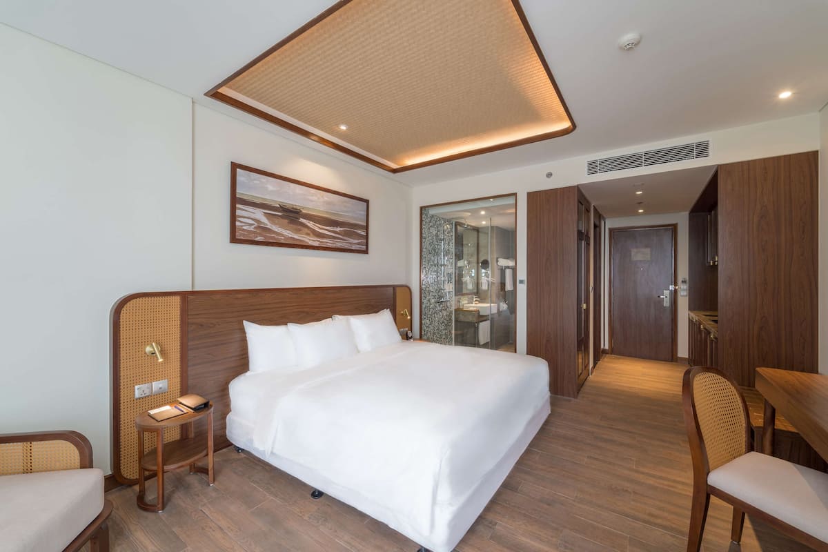 Ein Hotelzimmer mit einem großen Bett, einem Nachttisch, einem Sessel, einem Schreibtisch und einem durch eine Glastür einsehbaren Badezimmer.