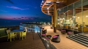 Un espace de détente extérieur moderne, doté d’une terrasse en bois, de sièges et d’une barrière en verre offrant une vue sur une étendue d’eau.