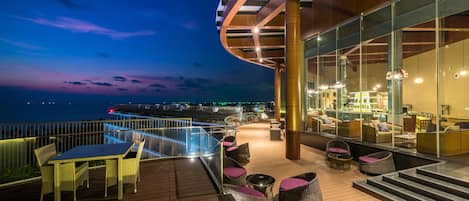 Un área lounge moderna al aire libre con terraza de madera, asientos y una barandilla de vidrio con vista a un cuerpo de agua.