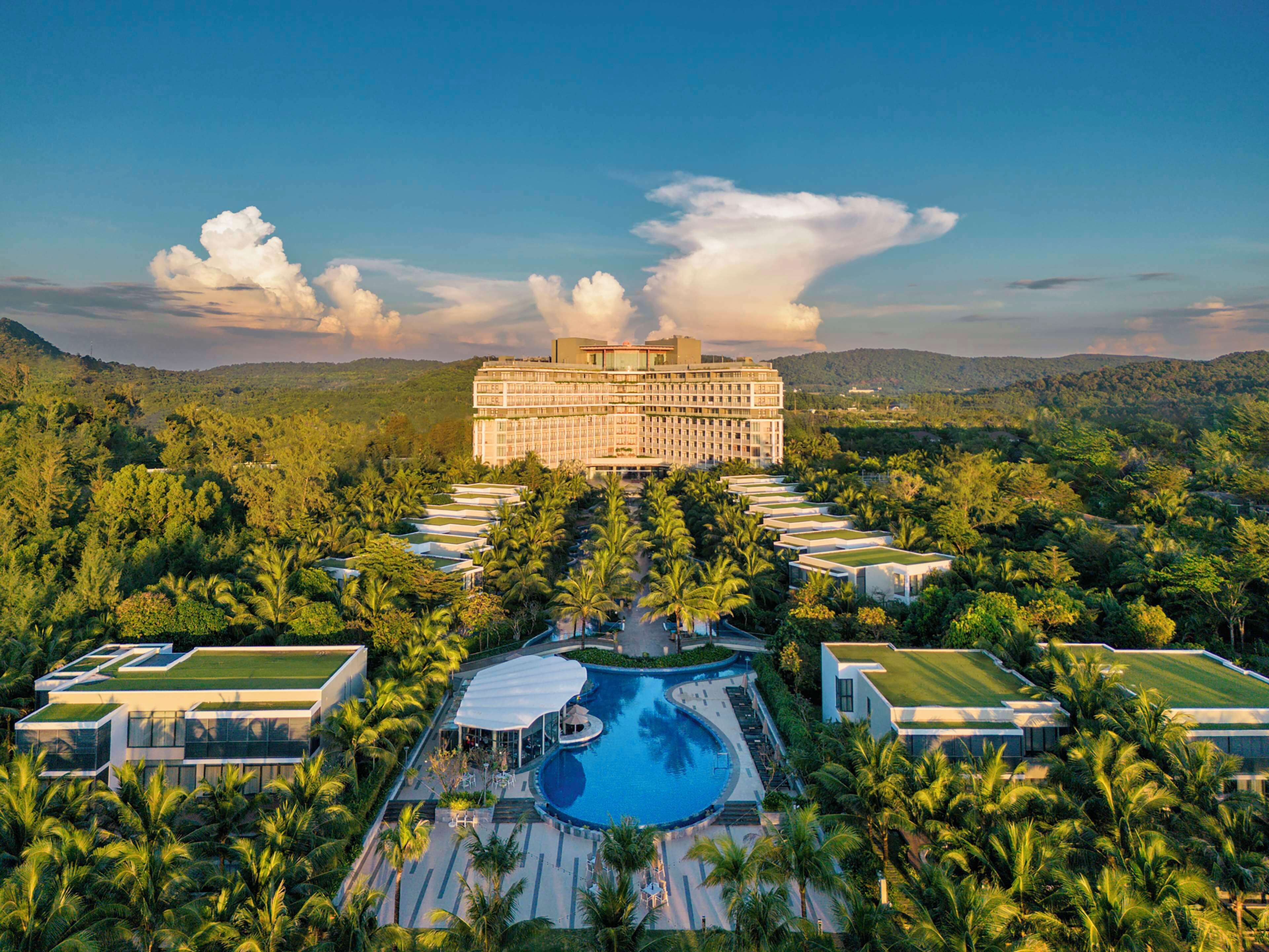 Ein großes Hotel mit einem zentral gelegenen Schwimmbecken, umgeben von üppiger Vegetation und Palmen.