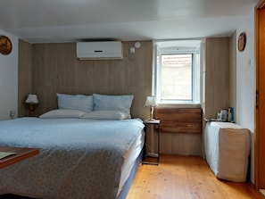 Un dormitorio con cama, lámparas de noche, una ventana, aire acondicionado montado en la pared y una mesita con libros y una figurita.