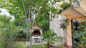 A patio area with a brick oven, a white table, and chairs, surrounded by greenery and a stone wall. - Apartment Cherry with spacious terrace & Wi-Fi (Krk)