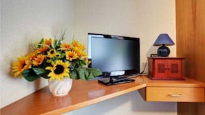 A television on a wooden desk with a vase of sunflowers and a small cabinet with a lamp. - Hotel Quellenhof (Bad Breisig)