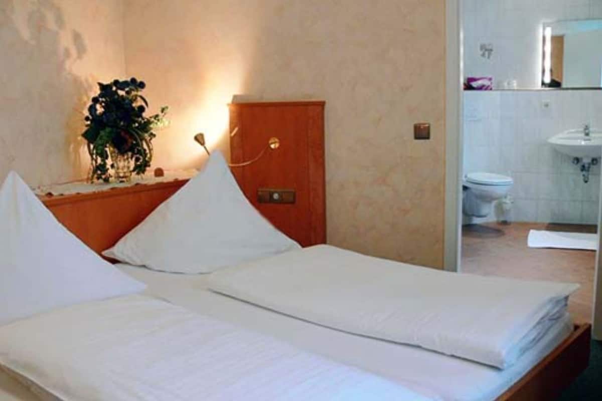 A hotel room with two beds, a wooden headboard, a bedside table with a plant, and a bathroom visible through a glass partition.