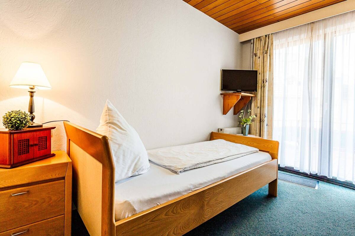 A single bed with a wooden frame, a bedside table with a lamp, a television mounted on the wall, and a window with curtains.