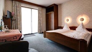 A hotel room with a wooden bed, a desk with a pink tablecloth, a television, and a large window with curtains. - Hotel Quellenhof (Bad Breisig)