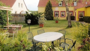 A patio with a round table and chairs, surrounded by greenery and a yellow building with a red roof.