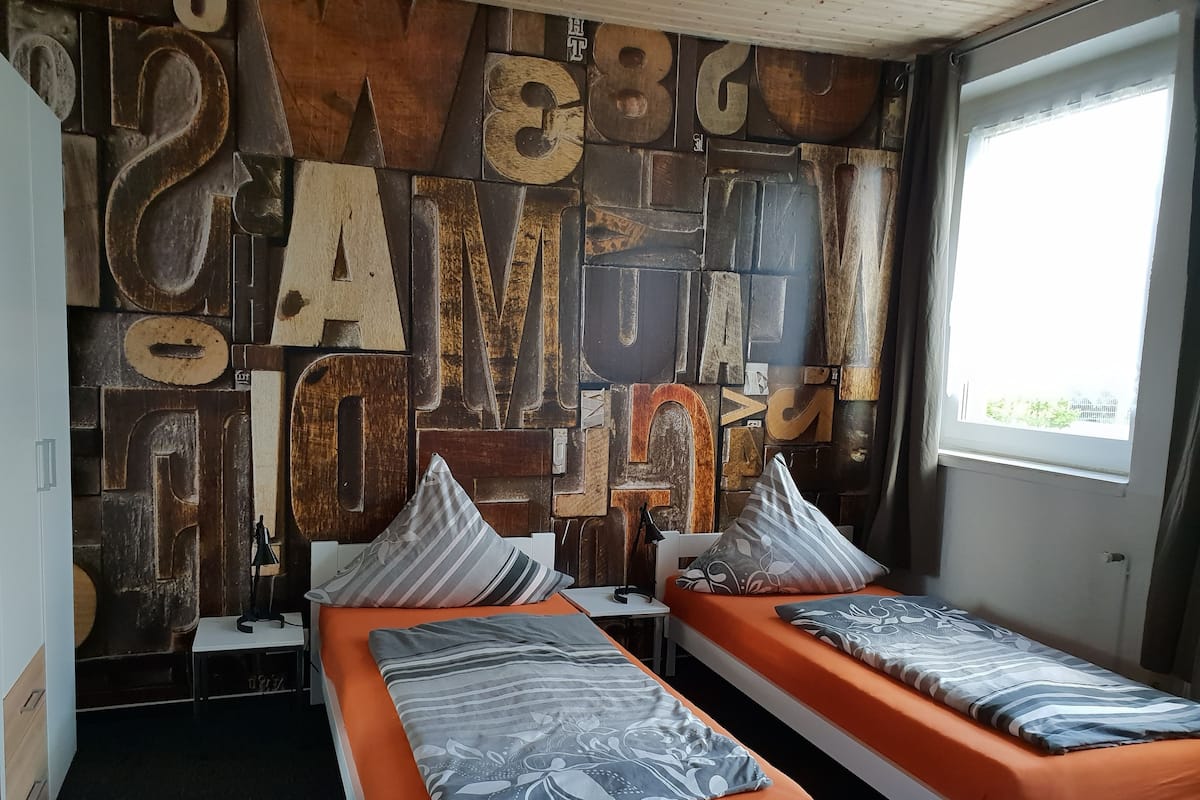 A room with two beds, each with an orange mattress and striped pillows. The walls are decorated with large, rusted letters.