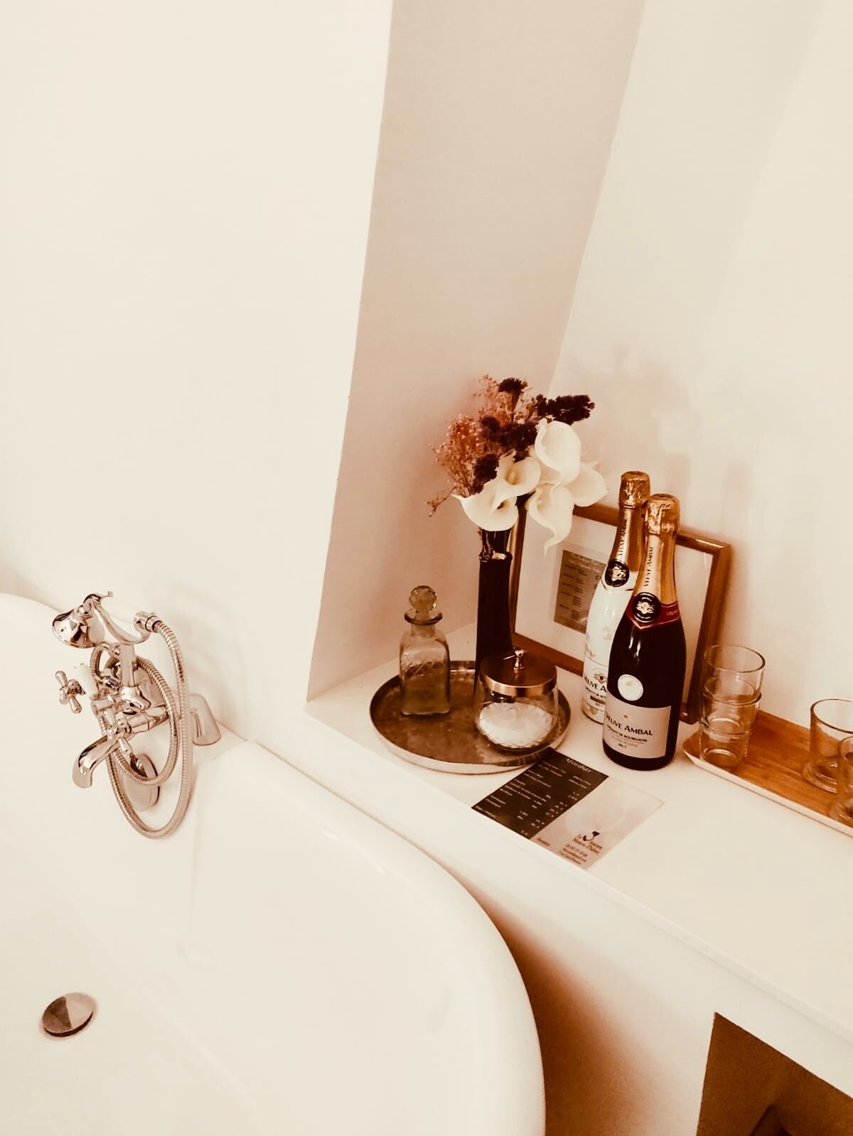 A bathroom with a bathtub, a vanity with a mirror, a bottle of champagne, and a vase with flowers.
